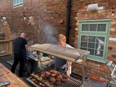 Two chefs grilling burgers and buns on a large outdoor grill against a brick wall adorned with string lights and a green window.