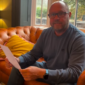 A man with glasses and a beard sits on a brown leather sofa, holding a white paper and smiling at the camera.