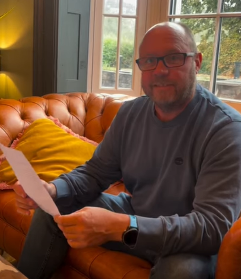 A man with glasses and a beard sits on a brown leather sofa, holding a white paper and smiling at the camera.
