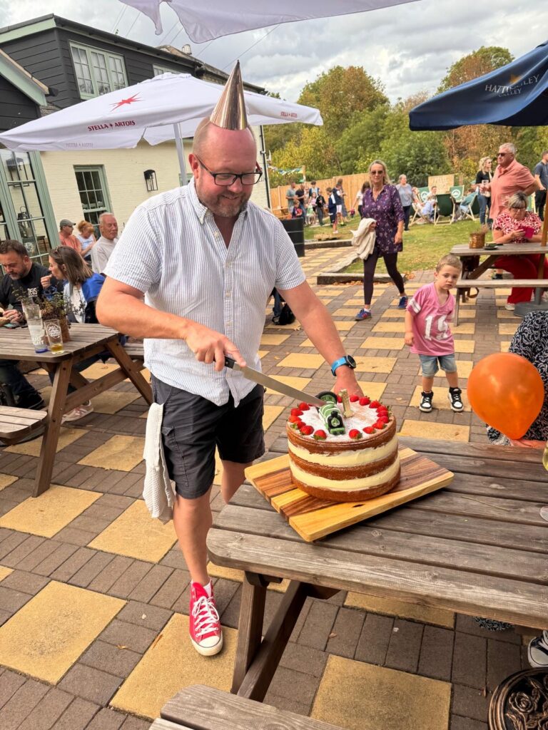 A smiling man in a party hat and glasses cuts a layered cake decorated with strawberries and beer bottles at an outdoor party.
