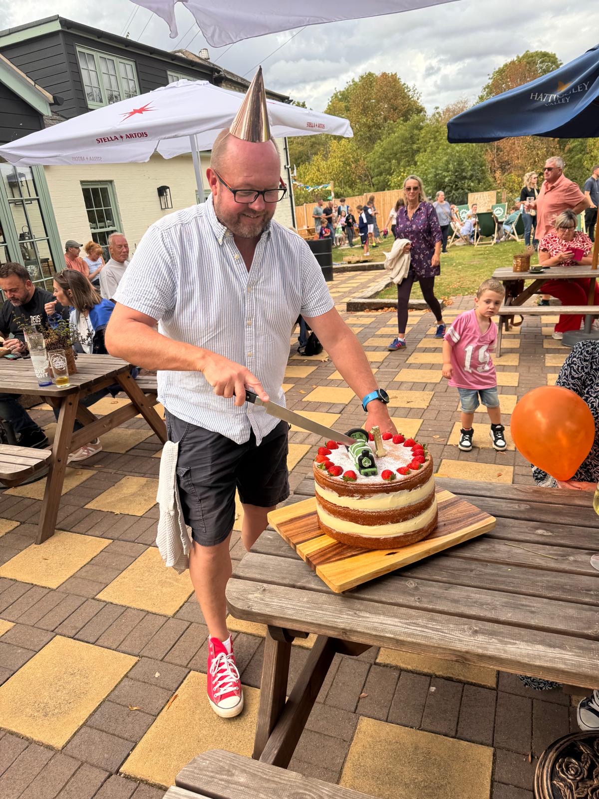 A smiling man in a party hat and glasses cuts a layered cake decorated with strawberries and beer bottles at an outdoor party.