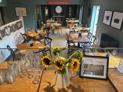 Interior view of a restaurant dining room with wooden tables, chairs, and dark green walls. Sunflowers in a pitcher are on a counter in the foreground.