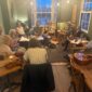 A group of adults gathered in a pub-like room with musical instruments, including guitars, violins, and a mandolin, for a jam session.