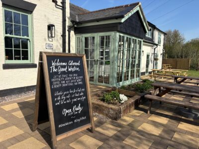 Exterior view of The Great Western pub on a sunny day, showing a chalkboard sign, a white building with green-framed windows and doors, and wooden picnic tables on a paved patio.