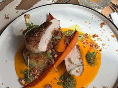 A gourmet meal on a white plate with sliced poultry, roasted carrots, orange purée, green pesto, and nuts. A bottle of Hildon sparkling water and a glass with an orange slice are in the background.