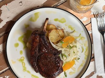 A meat chop with bone, roasted potatoes, and a fennel and orange salad on a white plate, with water and cutlery on a distressed wooden table.