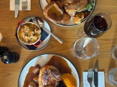 Overhead view of two roast dinners on a wooden table, featuring beef and chicken, with sides, wine, and cutlery.