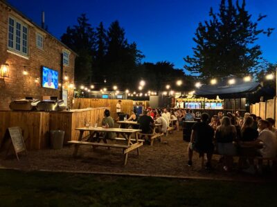 An outdoor pub garden at night, filled with people socializing at wooden picnic tables under string lights, with a TV screen on a brick wall and a bar in the background.