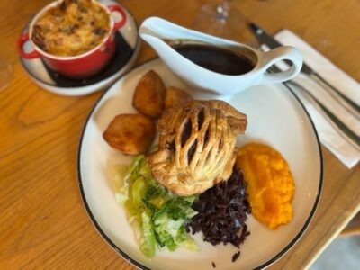 A full meal on a white plate with a pastry, roasted potatoes, green and red shredded vegetables, and orange mashed vegetables. A gravy boat and a small baked dish in a red ramekin are also visible.