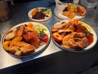 Three plates of roast dinners with beef, pork, potatoes, Yorkshire puddings, vegetables, and gravy on a metal counter.