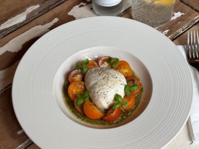 A white bowl containing burrata cheese, halved cherry tomatoes, green pesto, and basil leaves, on a wooden table.