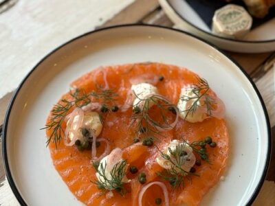 Smoked salmon dish with dill, capers, red onion, and cream cheese on a white plate, with bread and butter in the background.