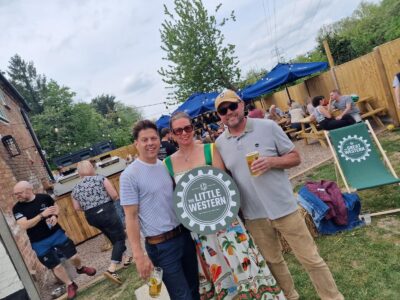 Three smiling people, two men and one woman, pose outdoors. The woman holds a round sign that says 'The Little Western'. All three are holding drinks. In the background are picnic tables, umbrellas, and other people.