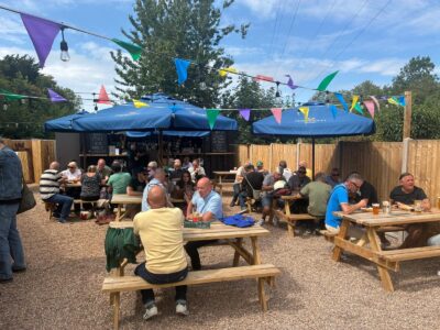 An outdoor pub garden with many people sitting at wooden picnic tables under blue umbrellas and colorful bunting on a sunny day.