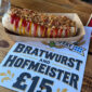 A large bratwurst in a bun topped with ketchup, mustard, and crispy onions, in a cardboard tray on a wooden table, next to a sign advertising 'Bratwurst and Hofmeister' for £15.