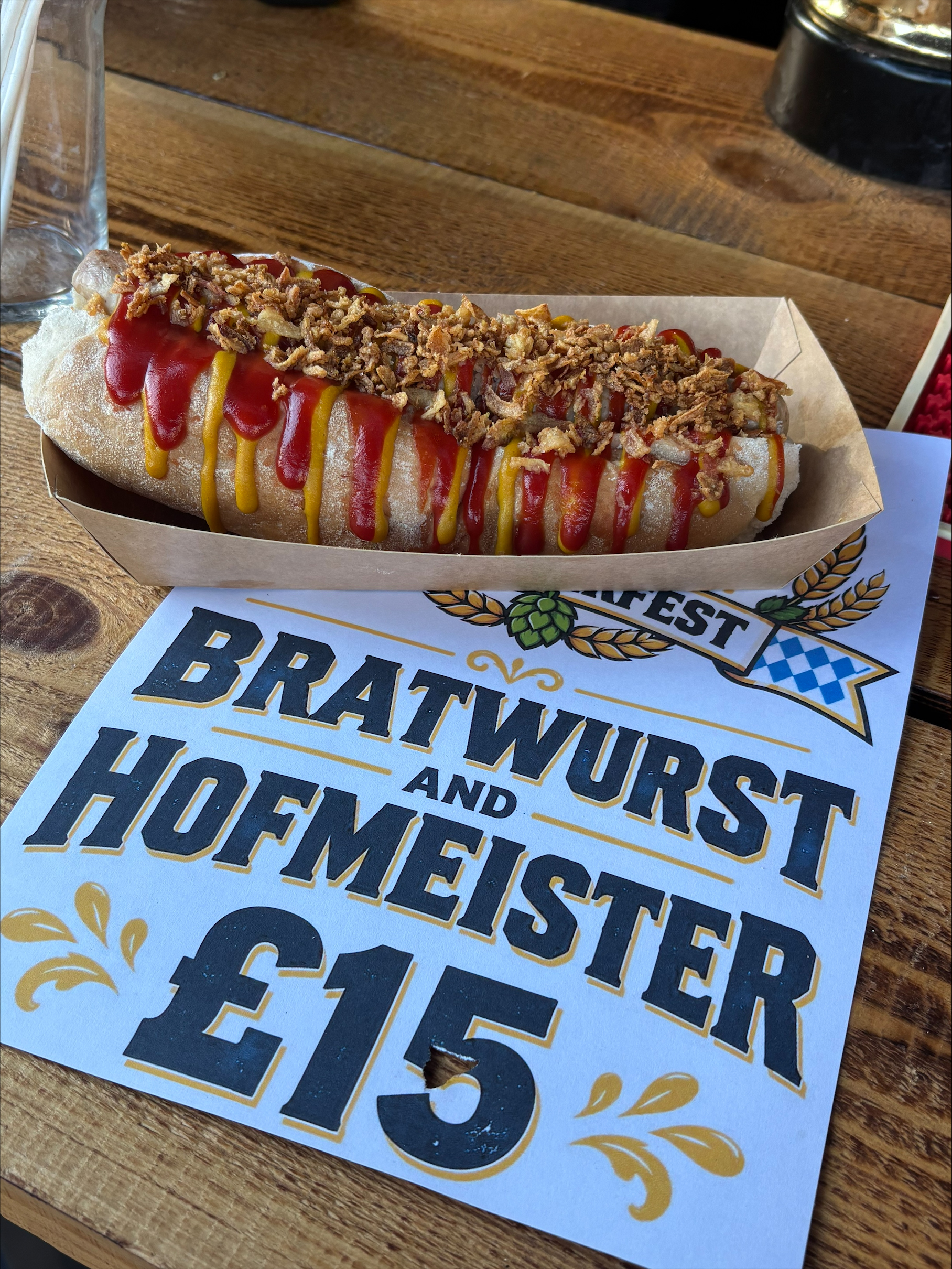 A large bratwurst in a bun topped with ketchup, mustard, and crispy onions, in a cardboard tray on a wooden table, next to a sign advertising 'Bratwurst and Hofmeister' for £15.