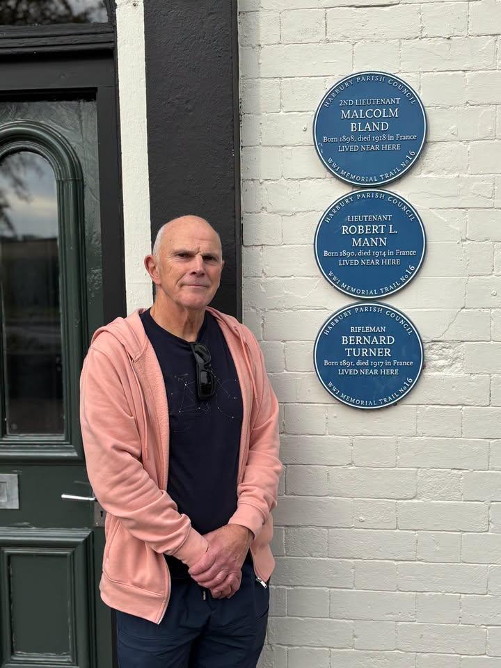 A bald man in a pink hoodie stands next to a white brick wall with three blue circular plaques honoring WWI soldiers.