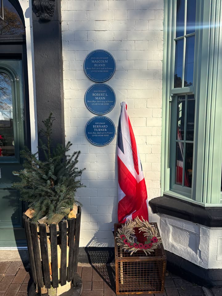 Three blue memorial plaques on a white brick wall, commemorating WW1 soldiers. A Union Jack flag and a poppy wreath sit below, next to an evergreen tree in a planter.